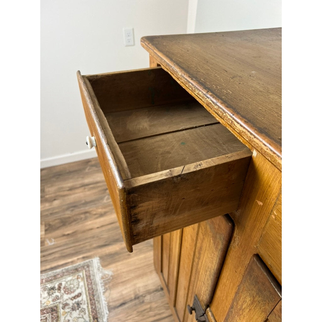 The Antique Victorian Country Grain Painted Jelly Cupboard c. 1870 stands on a wood floor by a patterned rug; its wooden drawer is pulled open, showing the empty, well-worn interior with a white wall outlet in the background.