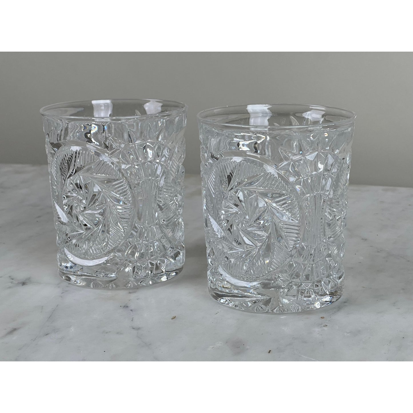 A pair of Fostoria Stowe Double Old Fashioned Tumbler Glasses with intricate cut crystal patterns sit side by side on a white marble surface against a plain light gray background.