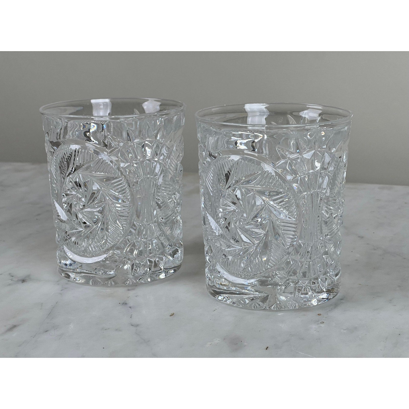 A pair of Fostoria Stowe Double Old Fashioned Tumbler Glasses with intricate cut crystal patterns sit side by side on a white marble surface against a plain light gray background.