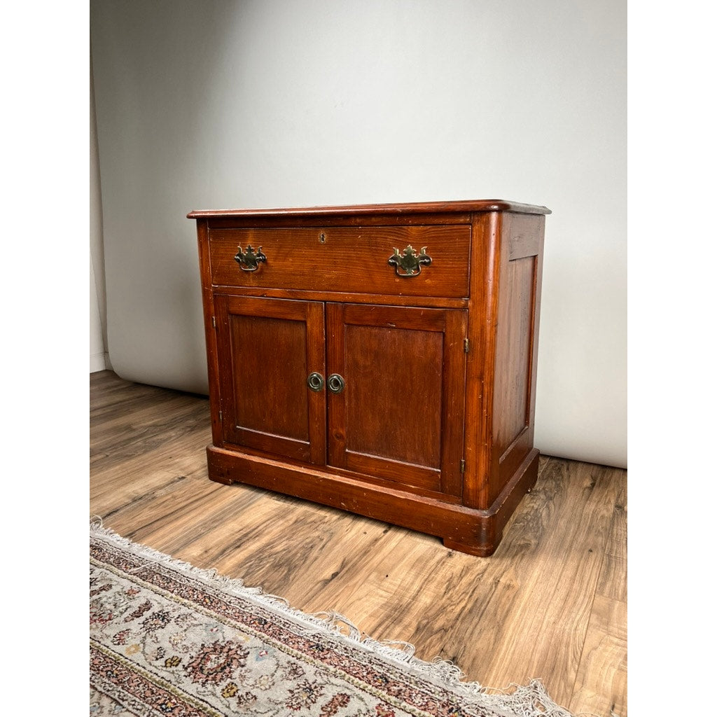 The Antique Victorian Pine Commode c. 1900 features a top drawer, two cabinet doors, and brass handles. It stands on a hardwood floor by a patterned rug against a light-colored wall. Brand: None and/or Unknown.