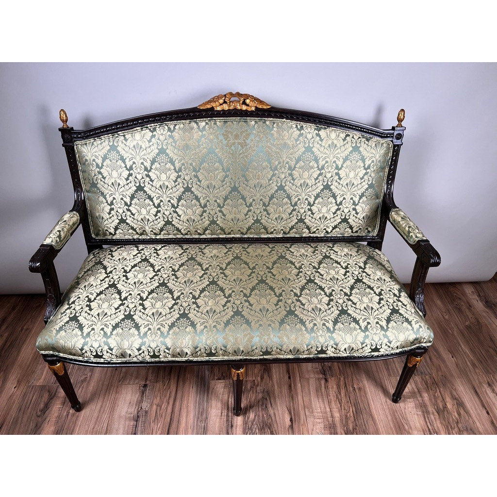 A Vintage Louis XVI Style Settee in green damask upholstery, featuring ornate carvings and gold accents, stands on a wooden floor against a plain gray wall. Brand: None/Unknown.