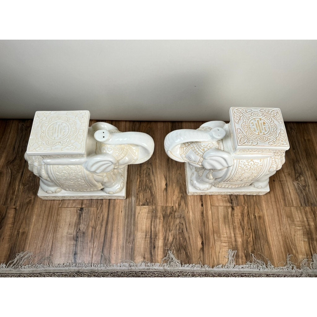 A vintage pair of white-on-white glazed Hollywood Regency style elephant garden seats sits side by side on a wooden floor next to a fringed rug, adding classic charm as unique accent tables.