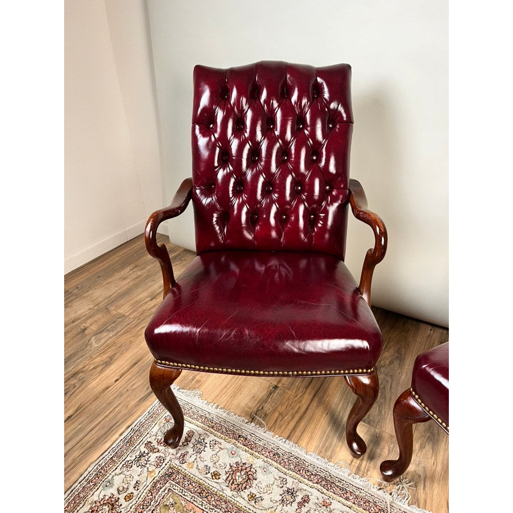 A Pair of Chippendale Style Tufted Burgundy Leather Library Armchairs sits on a wood floor next to a patterned rug, bringing timeless elegance into the space.