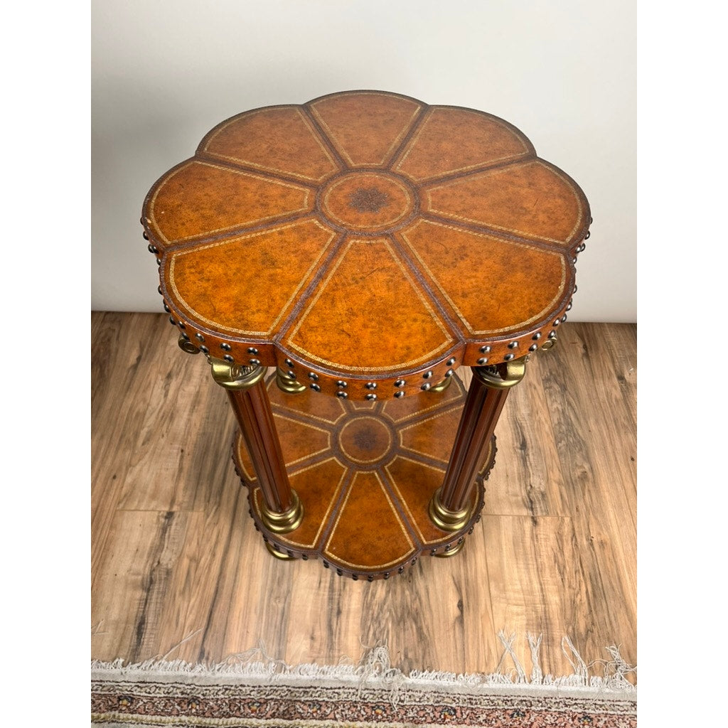 The Vintage Maitland-Smith Leather-Clad Occasional Table by Maitland Smith features a flower shape, brass nailhead trim, decorative brass accents, and two tiers, shown on a wooden floor beside a patterned rug.