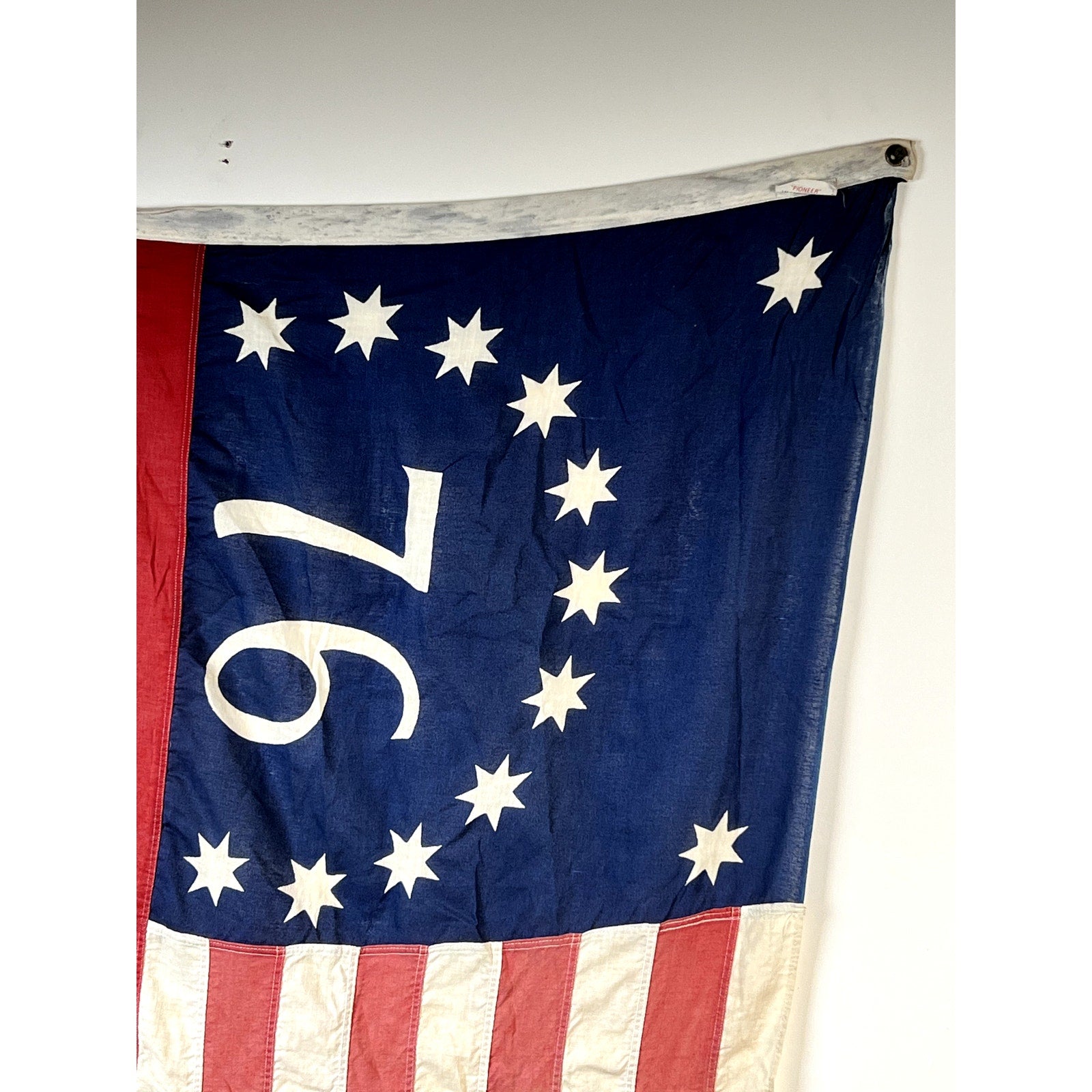 A section of a Vintage Valley Forge “76” Bennington American Flag from the Bicentennial Era (c. 1976), featuring the iconic “76,” thirteen stars arranged in a circle on a blue canton, and bold red and white stripes along the bottom, is displayed against a plain white wall.