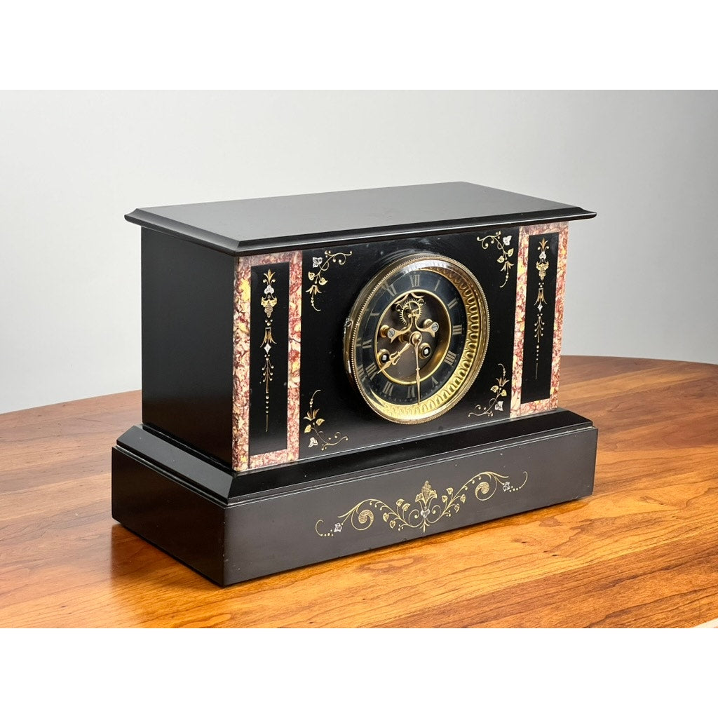 An Antique Victorian Slate Mantle Clock with Open Escapement, 8 Day Time & Strike (late 19th c.), featuring Roman numerals and ornate floral details, sits on a wooden surface against a plain background. Brand unknown.