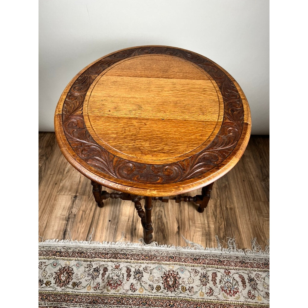 Antique English Oak Carved Gateleg Occasional Table c. Late 19th Century with natural wood grain and a decorative circular carved border, shown on a wooden floor beside a fringed patterned rug. Brand: None/Unknown.