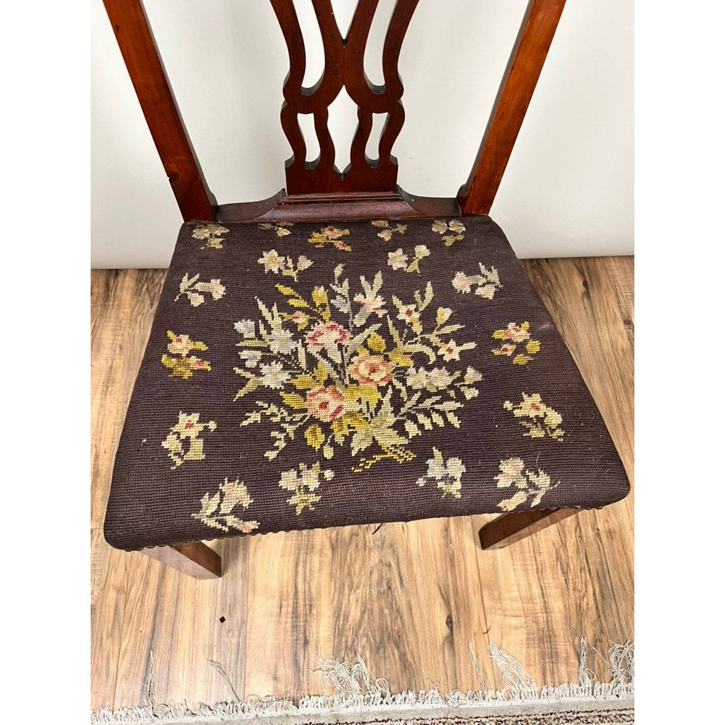 An Antique Chippendale Side Chair with Needlepoint Seat c. Mid 19th Century features a pierced splat back and dark floral-patterned seat with yellow, white, and pink flowers. It sits on a wood floor beside a light fringe rug. Brand: Unknown.