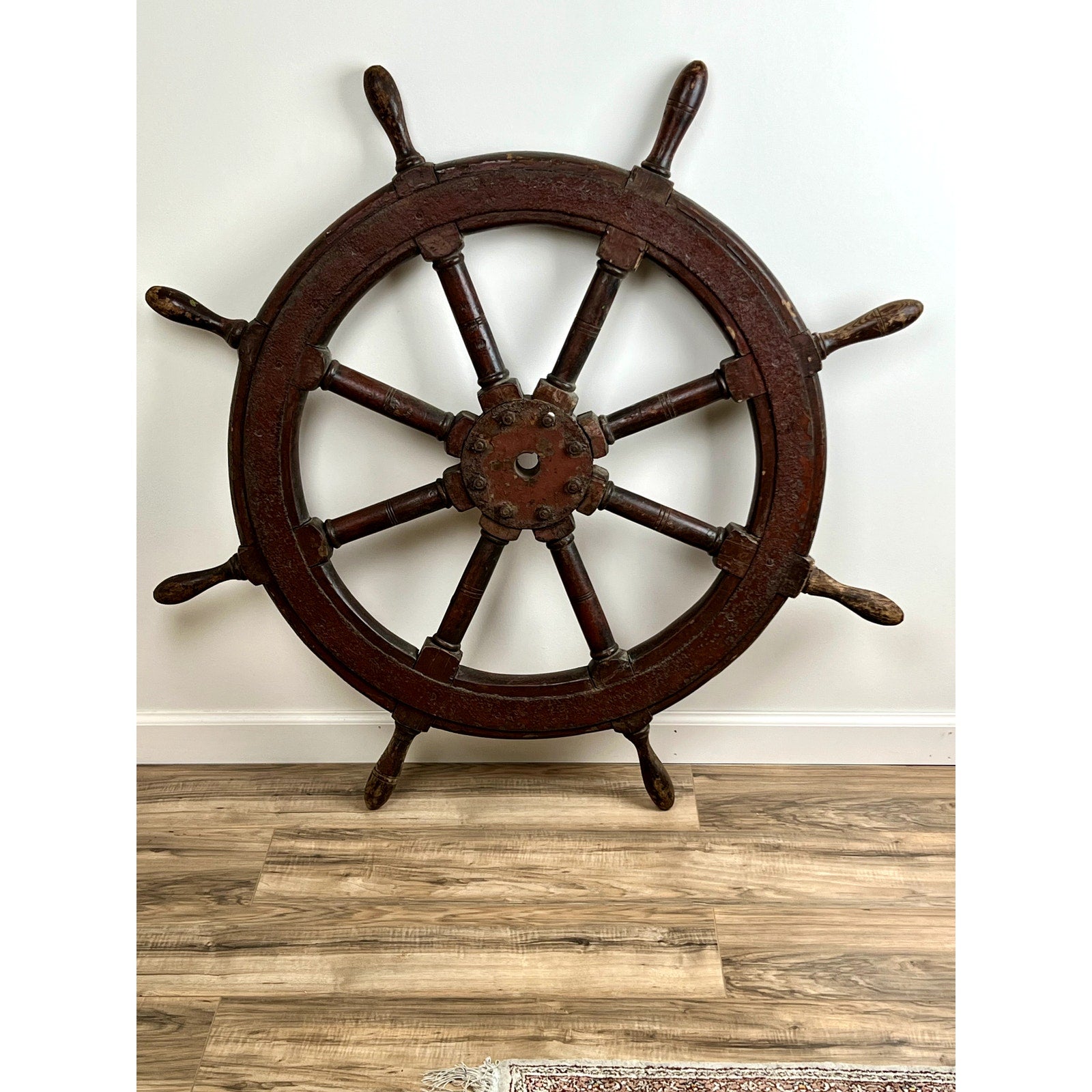 An antique eight spoke mahogany and iron ship’s wheel, stamped “1864 US,” leans against a white wall on a wood floor. Its dark, worn finish adds authentic 19th-century nautical charm; part of a rug is visible in the lower right corner.