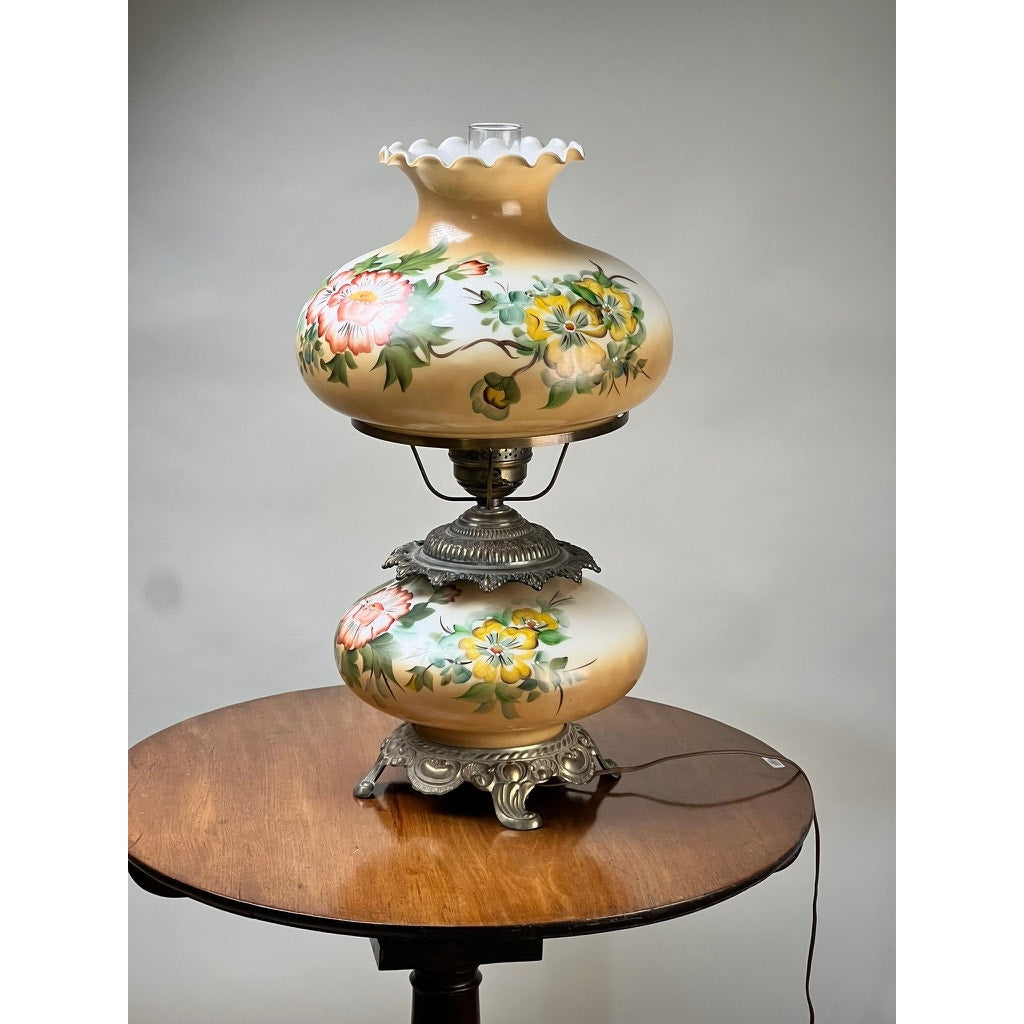 A Vintage Hand-Painted Floral Milk Glass Hurricane Parlor Lamp with ornate metal detailing and a visible power cord sits on a round wooden table, showcasing classic charm. Brand: None/Unknown.