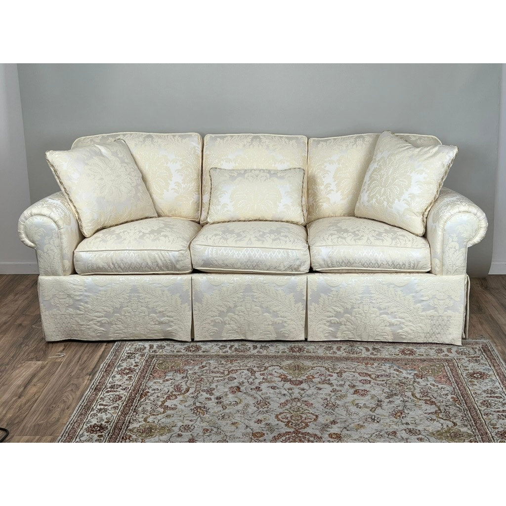 A Vintage Custom Ivory Upholstered Sofa by Stickley with three cushions and rolled arms sits on a wood floor in front of a light gray wall, with a floral area rug placed in front.
