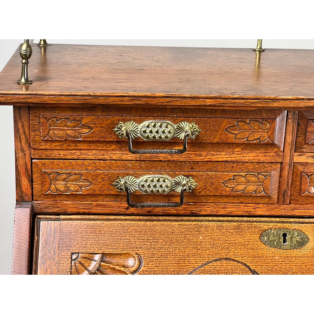 Close-up of an Antique Victorian Oak Ladies Writing Desk c. 1900, featuring ornate brass handles, carved leaf patterns, and a warm oak finish with a visible keyhole on the lower cabinet. Brand: Unknown.