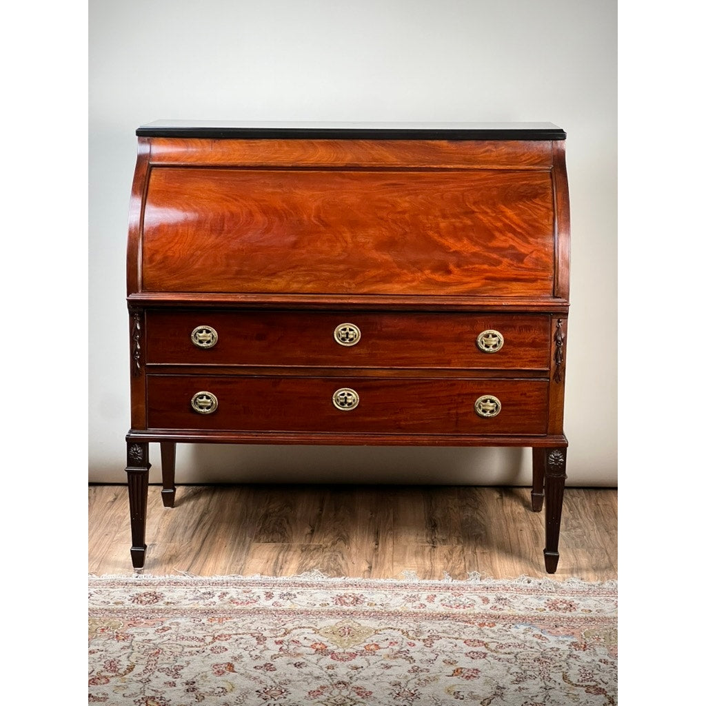 An Antique Directoire Style French Cylinder Desk with Slate Top, c. Late 19th Century, stands on a patterned rug against a neutral wall. This desk features a curved roll-top, two large drawers with brass handles, and elegant tapered legs.