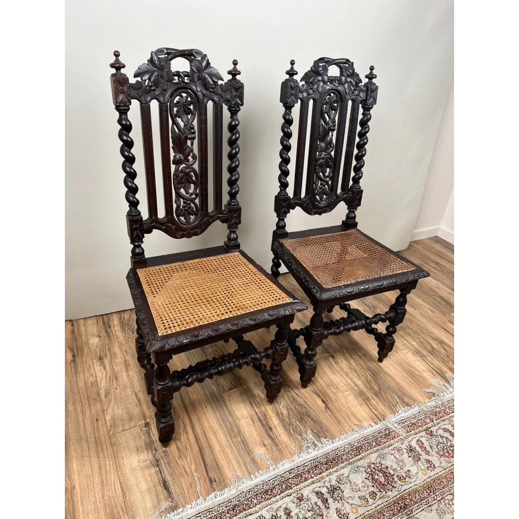 A Pair Antique French Walnut Hunting Chairs c. Late 19th Century, with carved backs, twisted legs, and woven cane seats (one damaged), are side by side on a wood floor, with a patterned rug partially visible in the foreground.