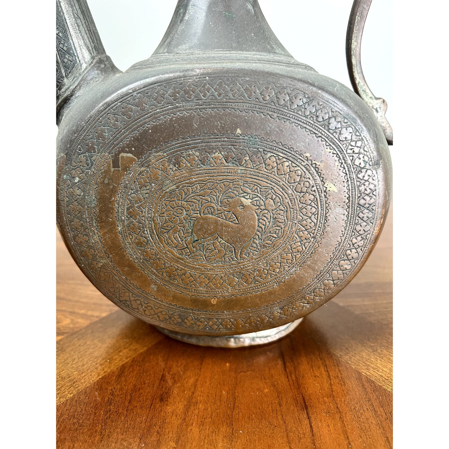 Close-up of an Antique Qajar Persian Copper Engraved Ewer c. 19th Century, featuring intricate patterns and a central deer with floral motifs, showcases exquisite Islamic-inspired Persian metalwork on a wooden surface. Brand: Unknown.