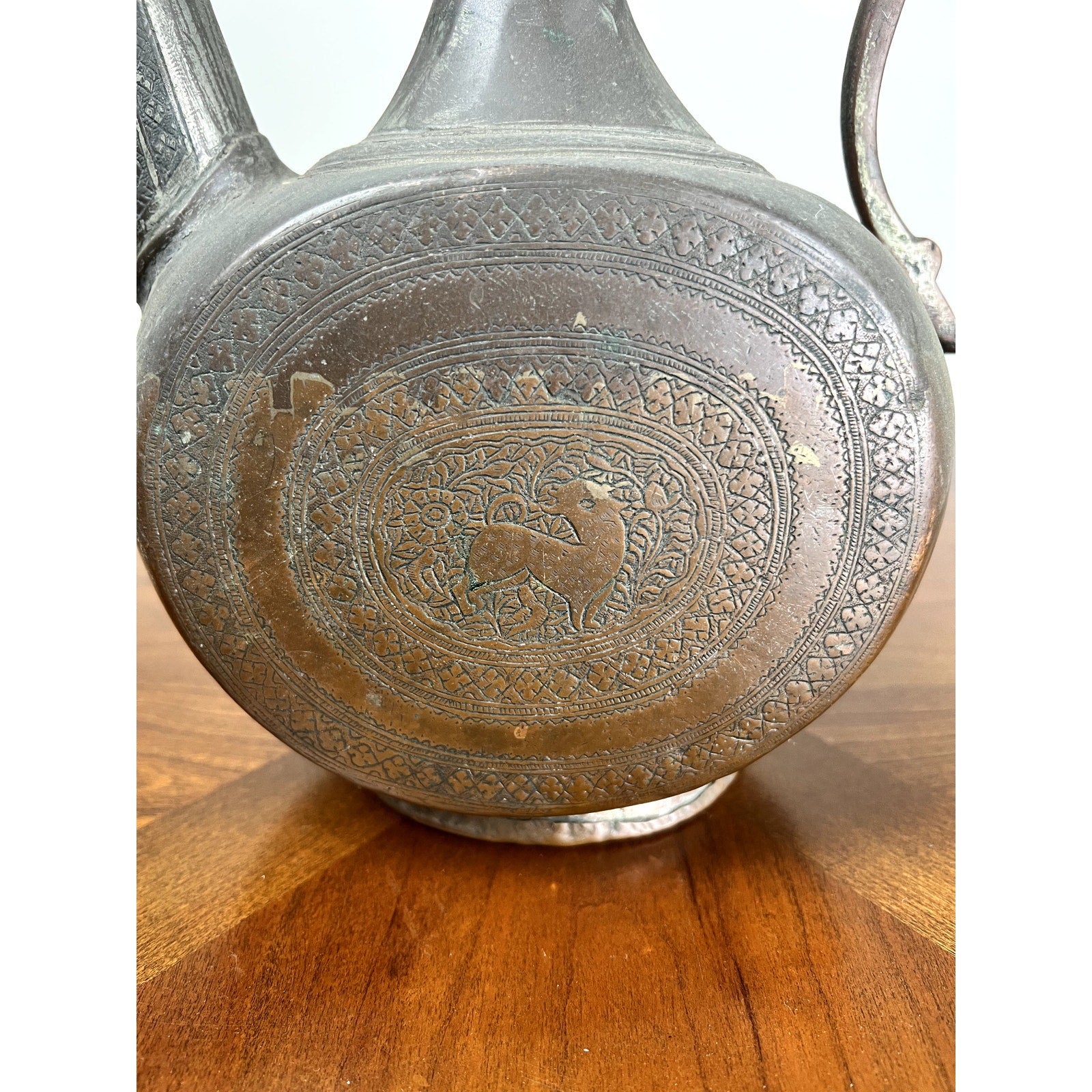 Close-up of an Antique Qajar Persian Copper Engraved Ewer c. 19th Century, featuring intricate patterns and a central deer with floral motifs, showcases exquisite Islamic-inspired Persian metalwork on a wooden surface. Brand: Unknown.