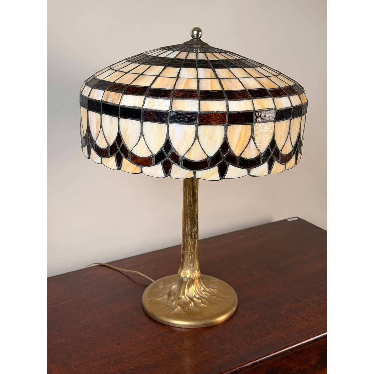 A Pearl City Antiques Vintage Tiffany Revival Leaded Stained Glass Table Lamp with Cast Brass Tree-Form Base rests on a wooden surface, showcasing its geometric cream, brown, and black stained glass shade. The bronze-finished cast brass base is crafted in a tree form, with a cord extending discreetly from the back.