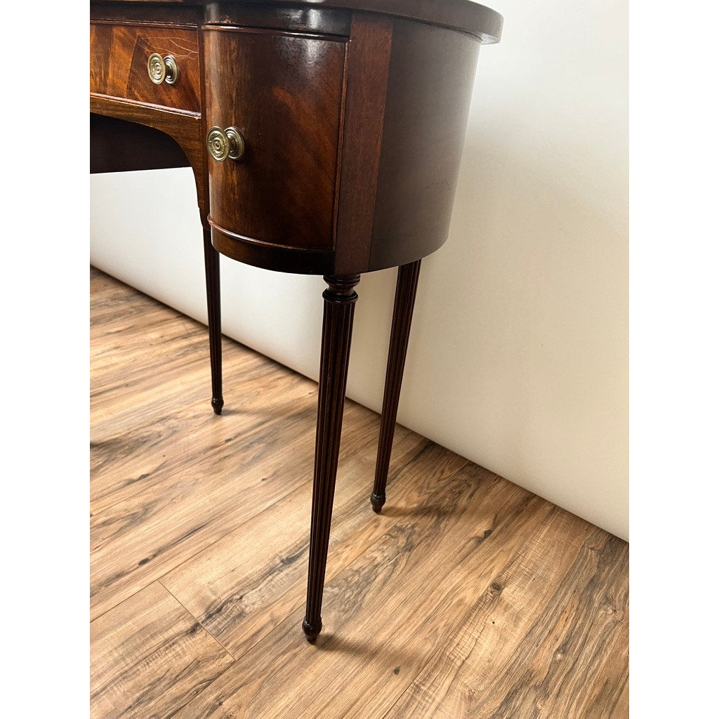 A close-up of a Vintage Edwardian Sheraton Revival Kidney-form Dressing Table c. 1940, with mahogany veneers, curved drawers, brass handles, and fluted legs, shown on a hardwood floor beside a white wall.