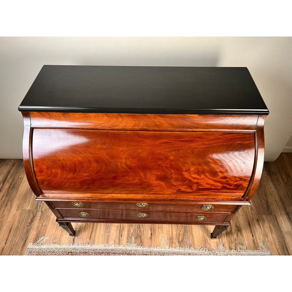 An Antique Directoire Style French Cylinder Desk with a slate top from the late 19th century stands on a wood floor, its curved roll-top front and brass drawer handles visible, with part of a rug at the bottom.