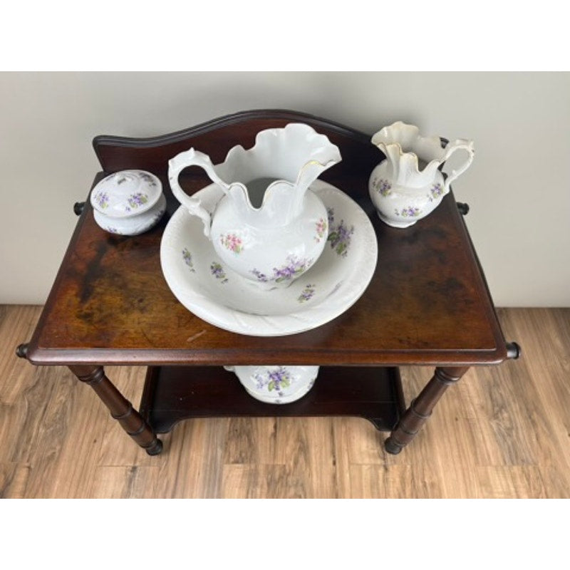A Leland Antique Victorian Porcelain Wash Stand Set (late 19th century) features white-and-purple floral pieces—lidded jar, matching pitchers, and extra bowl—displayed on a stand against a light wall and wood floor.