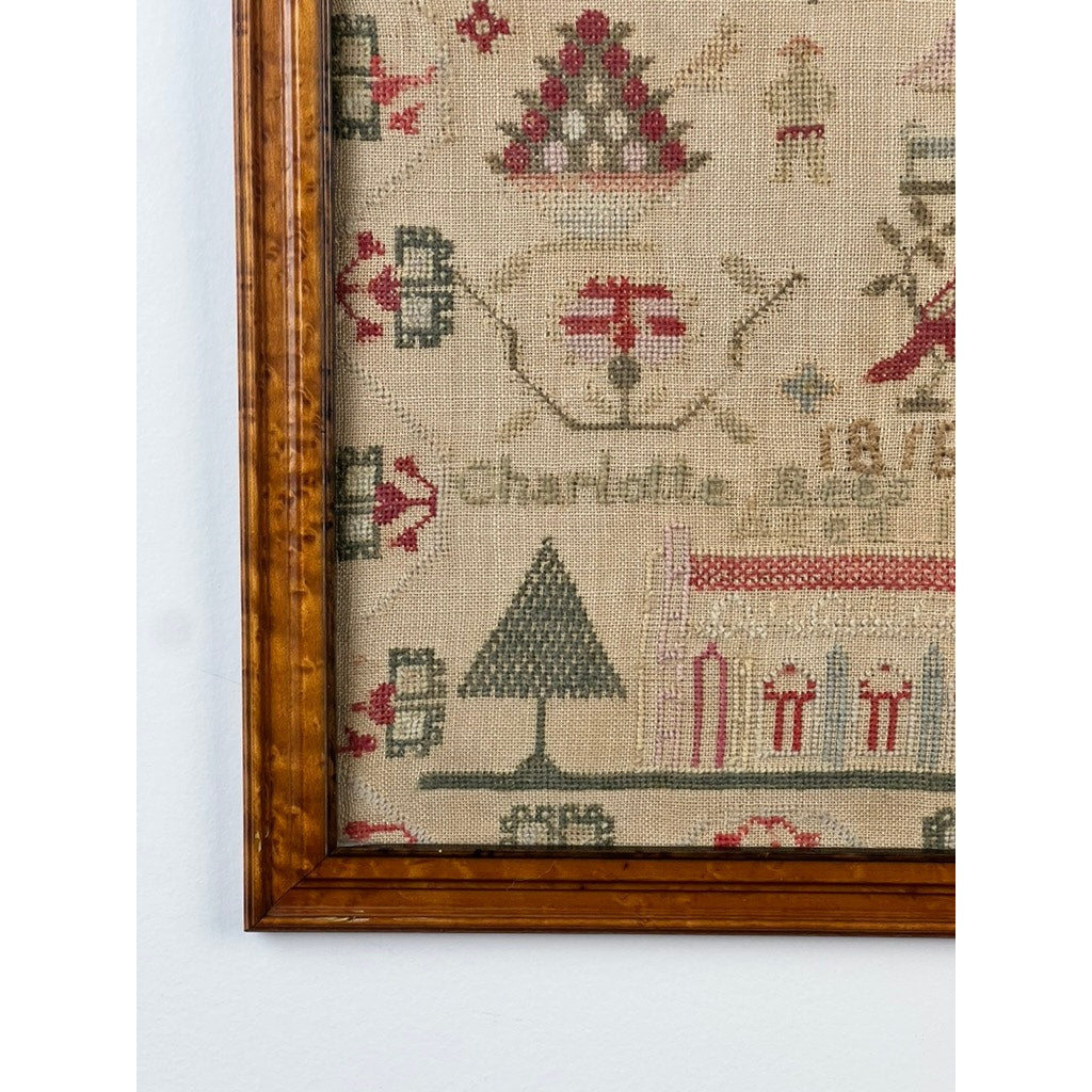 A close-up of the Exceptional English Regency Schoolgirl Needlework Sampler by Charlotte Rees, age 11 and dated 1816—showing intricate embroidery with a tree, geometric patterns, and stitched text on fabric, partially framed in wood against a white wall.