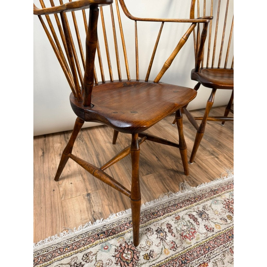 A close-up of a Pair of Signed L.E. Partridge Connecticut Bench-Made Continuous-Arm Windsor Chairs, c. 1960s, showcases the craftsmanship of these early American–style wooden chairs as they sit on a polished wood floor beside a patterned rug. Another matching chair is partially visible in the background, highlighting the timeless appeal of L.E. Partridge’s work.
