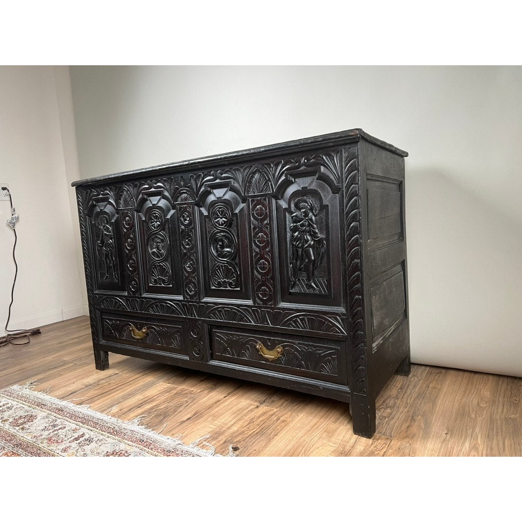 An Antique 18th Century Charles II Style Mule Chest, possibly Irish or Scottish, with carved panels, secret drawers, and brass-handled lower drawers stands on a wooden floor beside a patterned rug against a plain white wall.