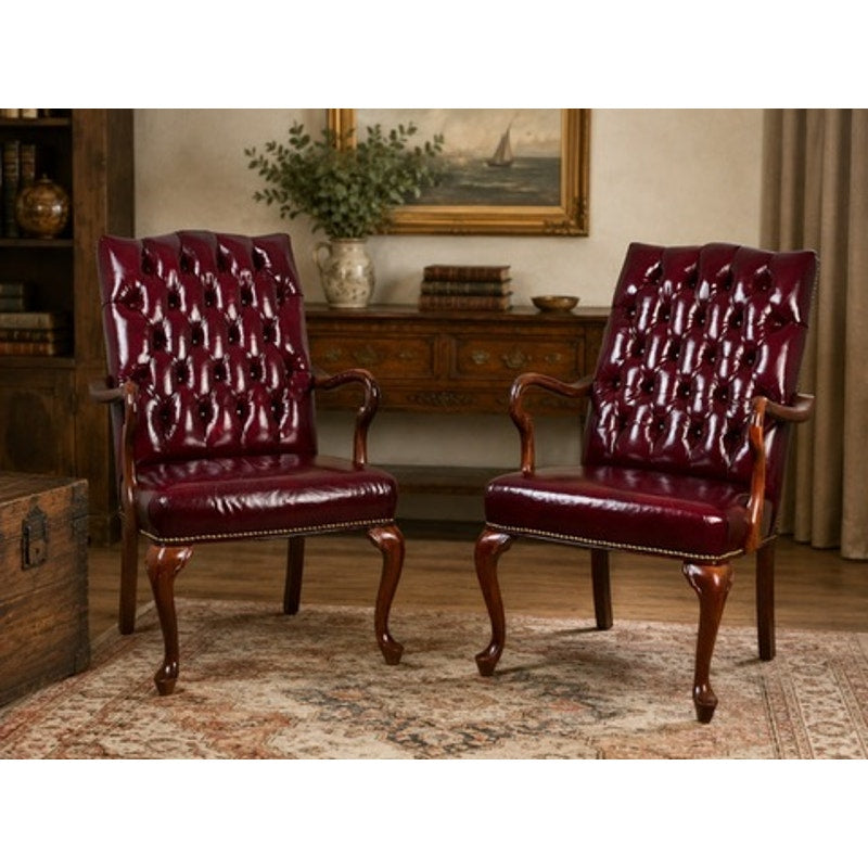 A pair of Chippendale Style Tufted Burgundy Leather Library Armchairs sit side by side on a patterned rug, in front of a wooden sideboard adorned with books, a vase of greenery, and a framed painting.