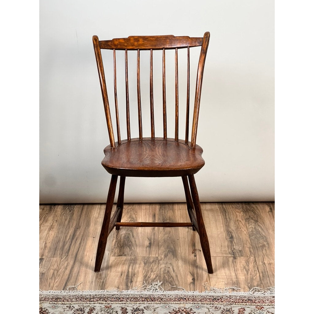 An Antique Step Down Windsor Chair in oak and pine, early 19th century, stands on a wooden floor with a portion of a patterned rug visible at the bottom. The chair features a curved backrest with vertical slats, contoured seat, and angled legs.