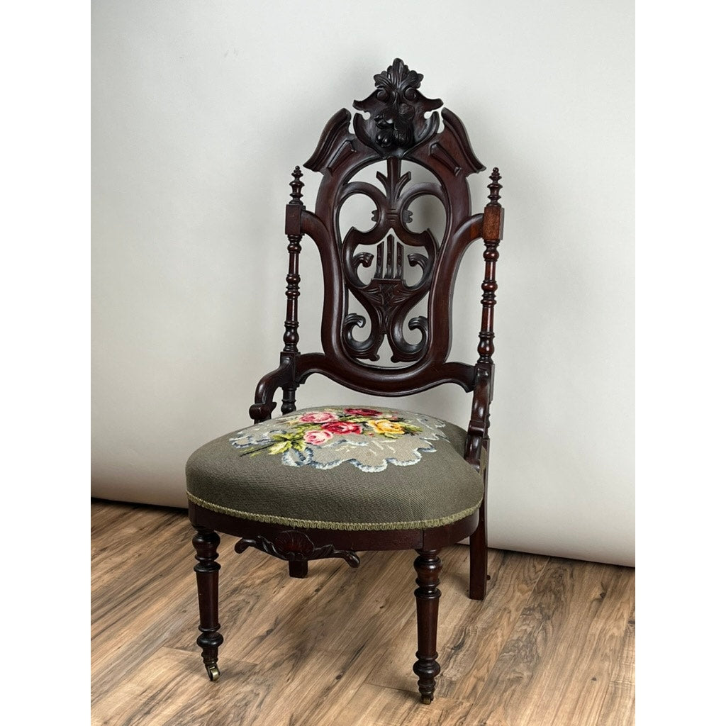 An Antique Victorian Rococo Revival Carved Walnut Slipper Chair with a floral needlepoint seat (c. late 19th century) features intricate carved details, exuding timeless elegance against a plain white wall and wooden floor. Brand: Unknown.