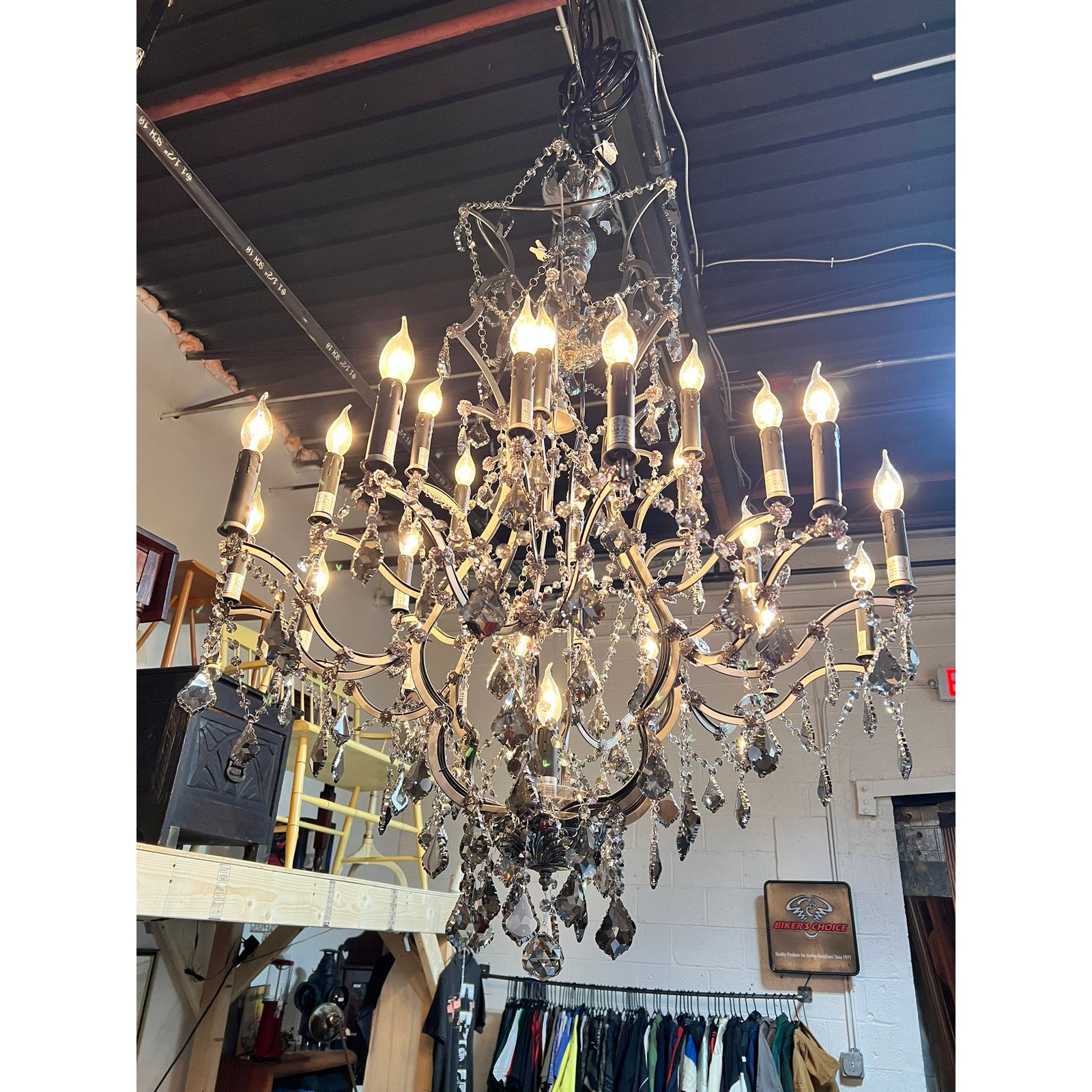 A Large Vintage Smoke Crystal 24 Light Chandelier hangs lit from a dark industrial ceiling, illuminating shelves, clothes racks, and assorted items in the background. Brand: None/Unknown.