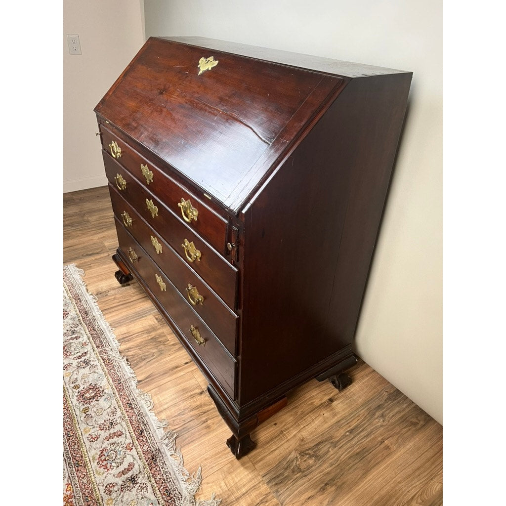 The 18th Century New England Chippendale Cherry Slant Front Desk, Circa 1770–1790, features a rich dark cherry wood finish, brass handles and keyholes, a classic slanted writing surface, and three spacious drawers. With a decorative rug partially visible on the hardwood floor beneath it, this antique piece beautifully reflects the craftsmanship of 18th-century New England.