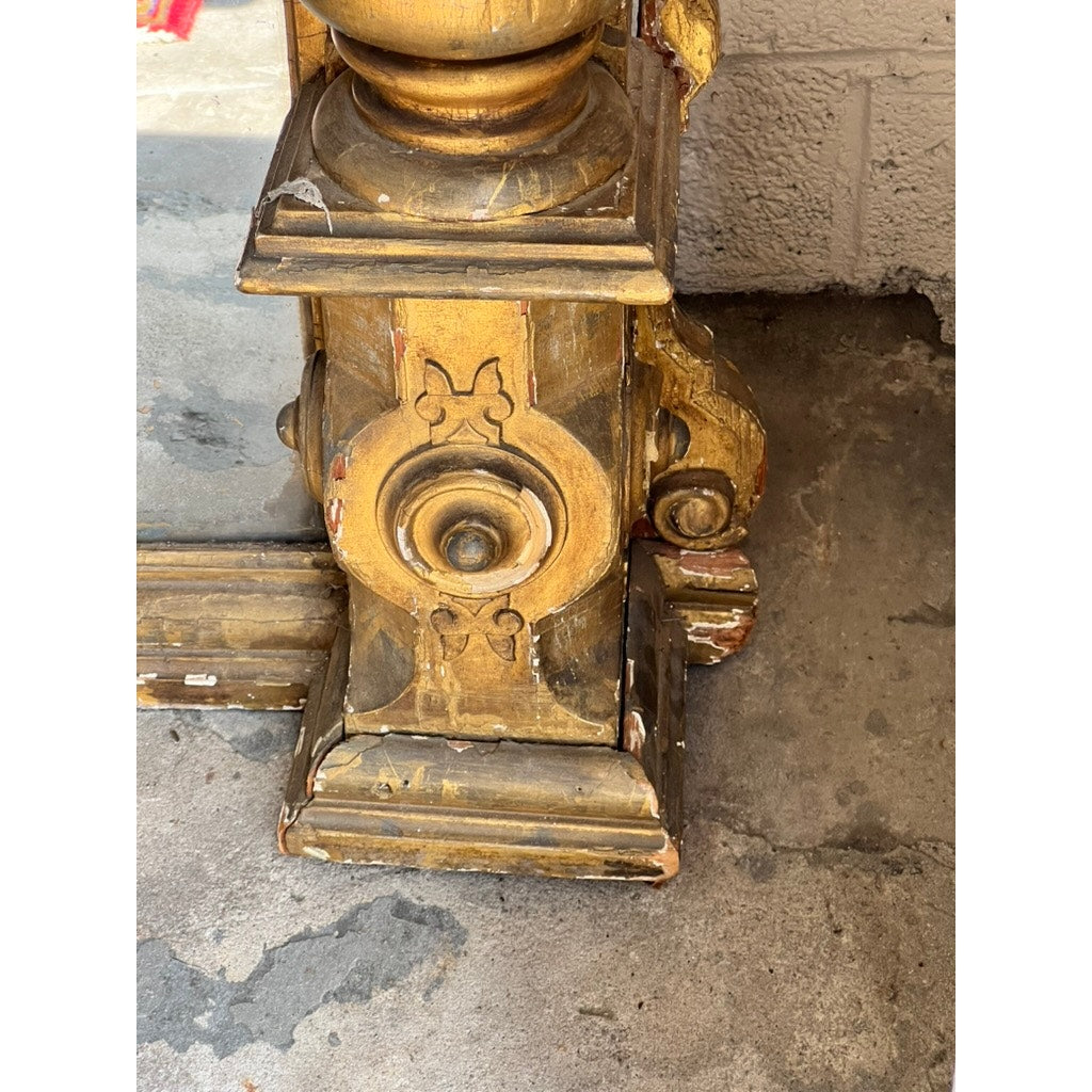 Close-up of an ornate, gold-painted wooden leg from an Exceptional Monumental Victorian Renaissance Revival Pier Mirror, Circa 1870–1885. The carved details include a butterfly motif, and the gilt paint is chipped to reveal underlying red and natural wood. The leg rests on a concrete floor beside a textured wall. Brand unknown.