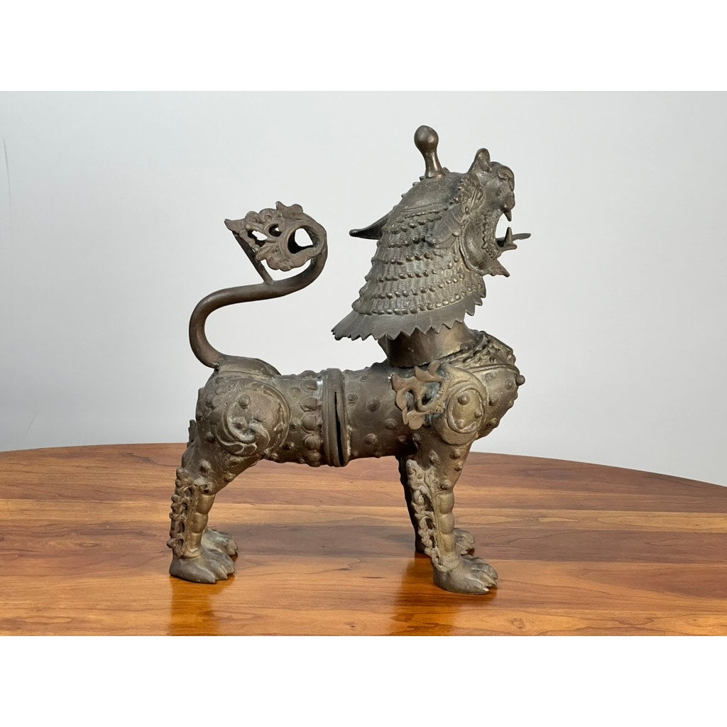 The Vintage 17” Cast Brass Pixiu Tibetan Lion c. 1960 features ornate detailing, a curled tail, and an open-mouthed head, standing on wood and reflecting Asian decorative elegance against a plain background. Brand: Unknown.