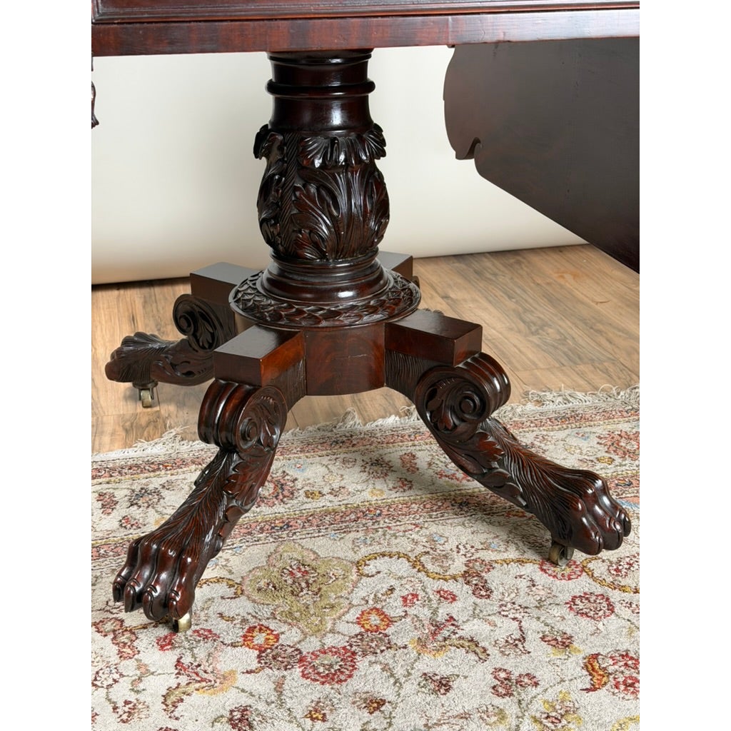 American Classical Carved Mahogany Drop-Leaf Breakfast Table, c. 1825–1840, featuring a dark wood pedestal with a carved central column and four legs ending in carved claw-and-ball feet with casters.