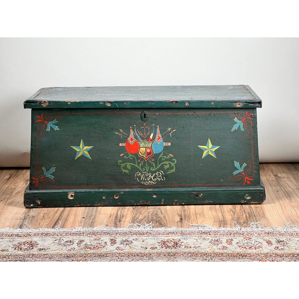 The Antique Paint Decorated English Sea Chest c. Late 19th Century features green paint, yellow stars, red and blue leaves, and a crest with red birds and shield. This vintage nautical trunk is displayed on wooden flooring by a patterned rug.