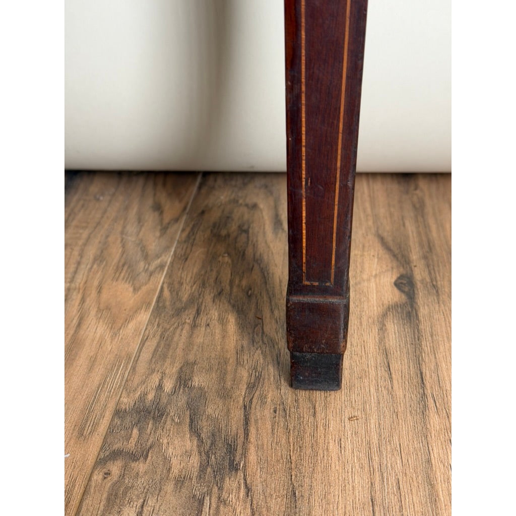 A tapered wooden leg with vertical light wood inlay from an English George III Mahogany and Satinwood-Inlaid Demilune Games Table, circa 1790–1810, resting on a wood floor.