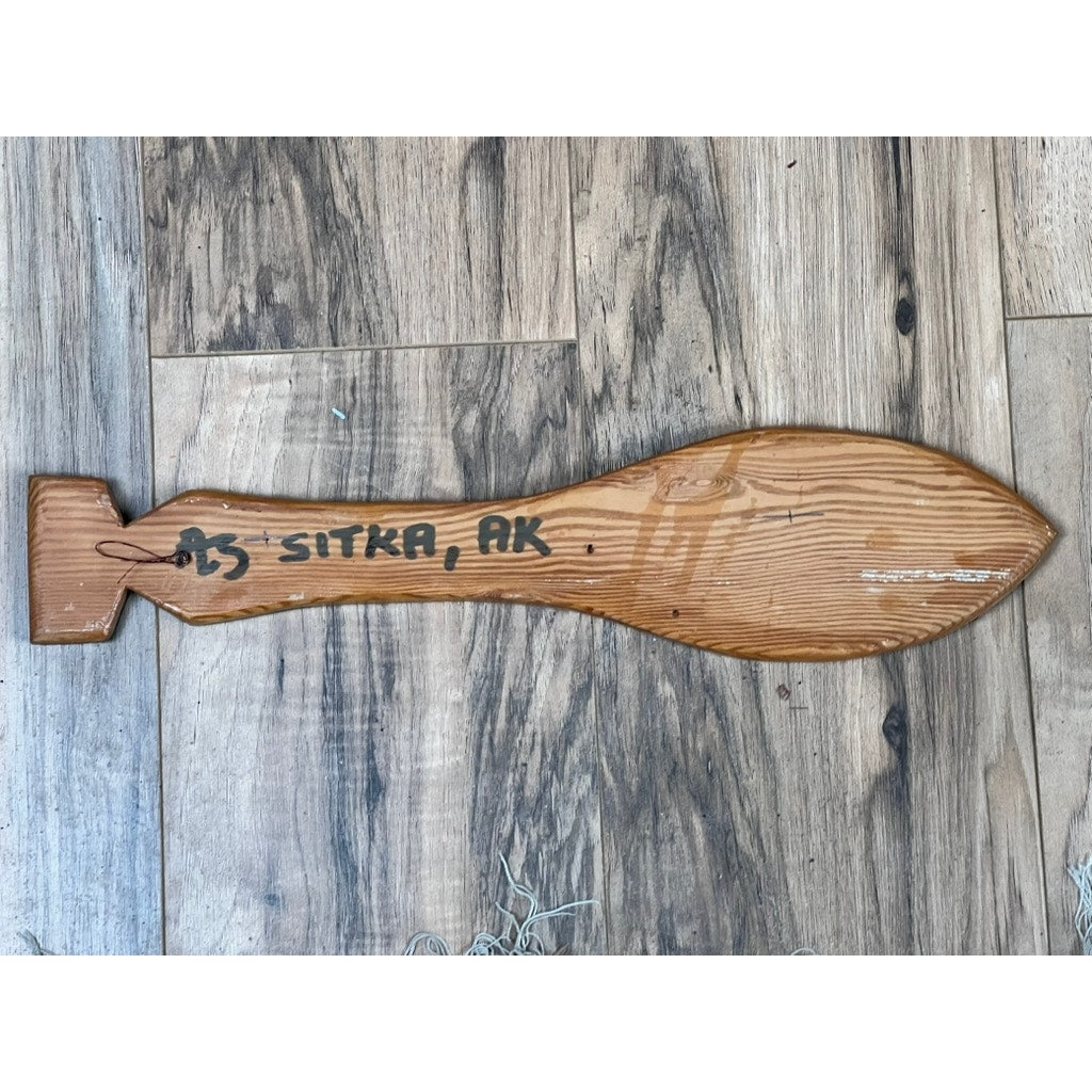 A Vintage Inuit Carved Red Cedar Paddle from Sitka, Alaska, signed illegibly, rests on a wood-patterned floor. Brand: None/Unknown.