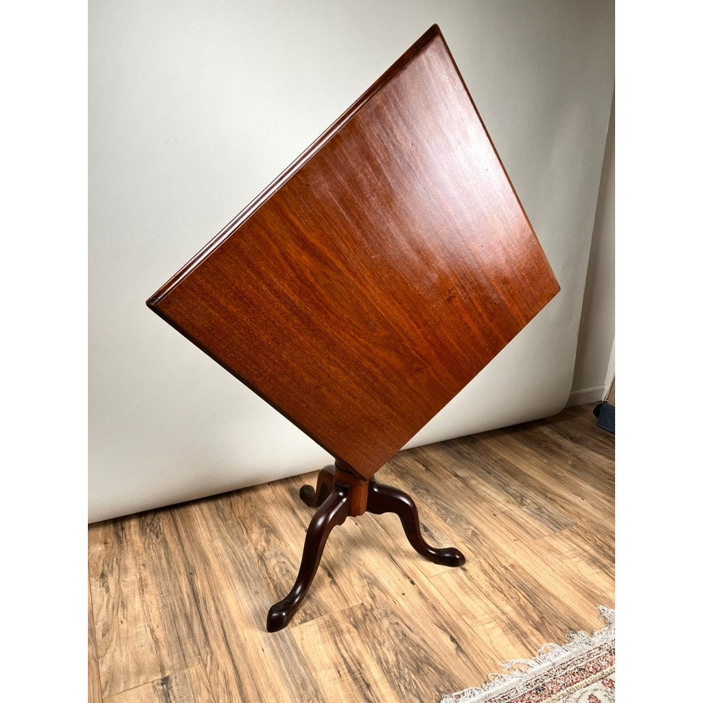 An Antique Queen Anne Mahogany Tilt Top Breakfast Table, c. Late 18th Century, stands on a wood floor with a plain background, featuring a polished square surface atop a central pedestal and three curved legs—an elegant historic piece.