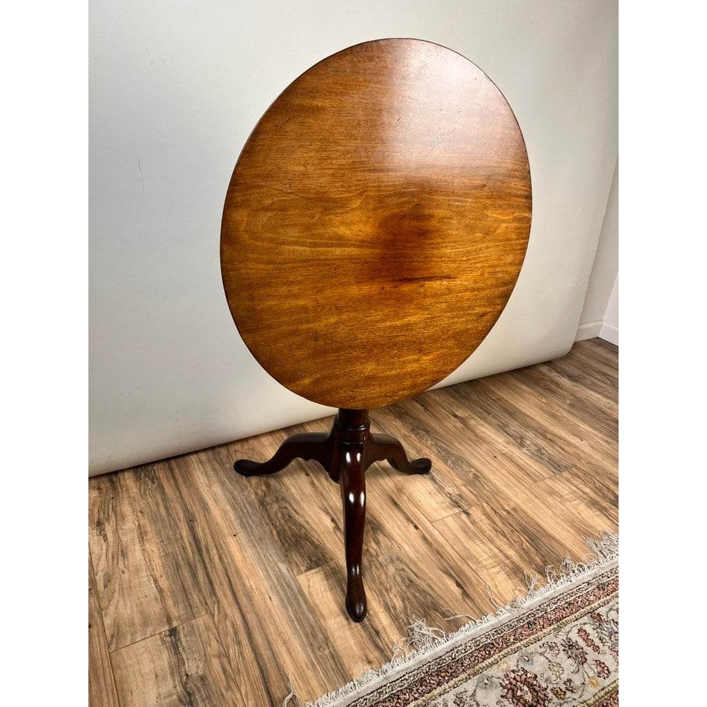 An Antique Queen Anne Mahogany Birdcage Tilt Top Breakfast Table (early 19th century) with a polished finish and three-legged pedestal base stands on a wooden floor near a patterned rug and light-colored wall.