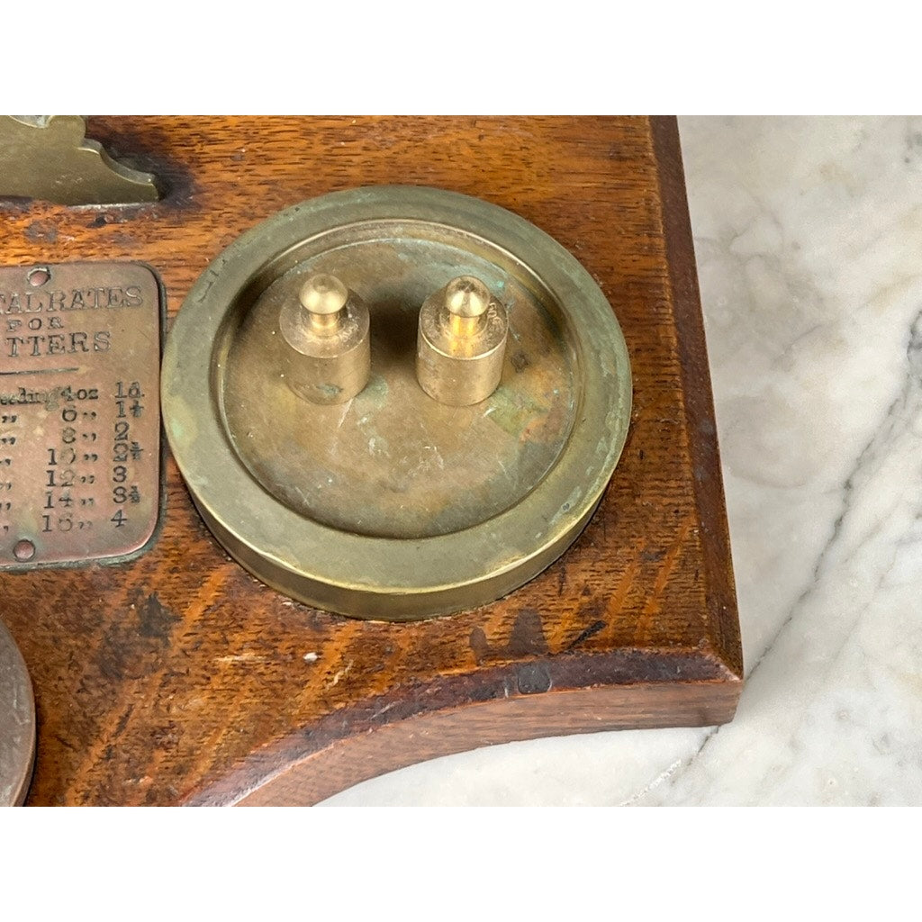 A close-up of the Antique 19th Century English Postal Scale's vintage wooden base, round brass component with two metal pegs, and engraved metal plaque, all displayed on a marble surface. Brand unknown.