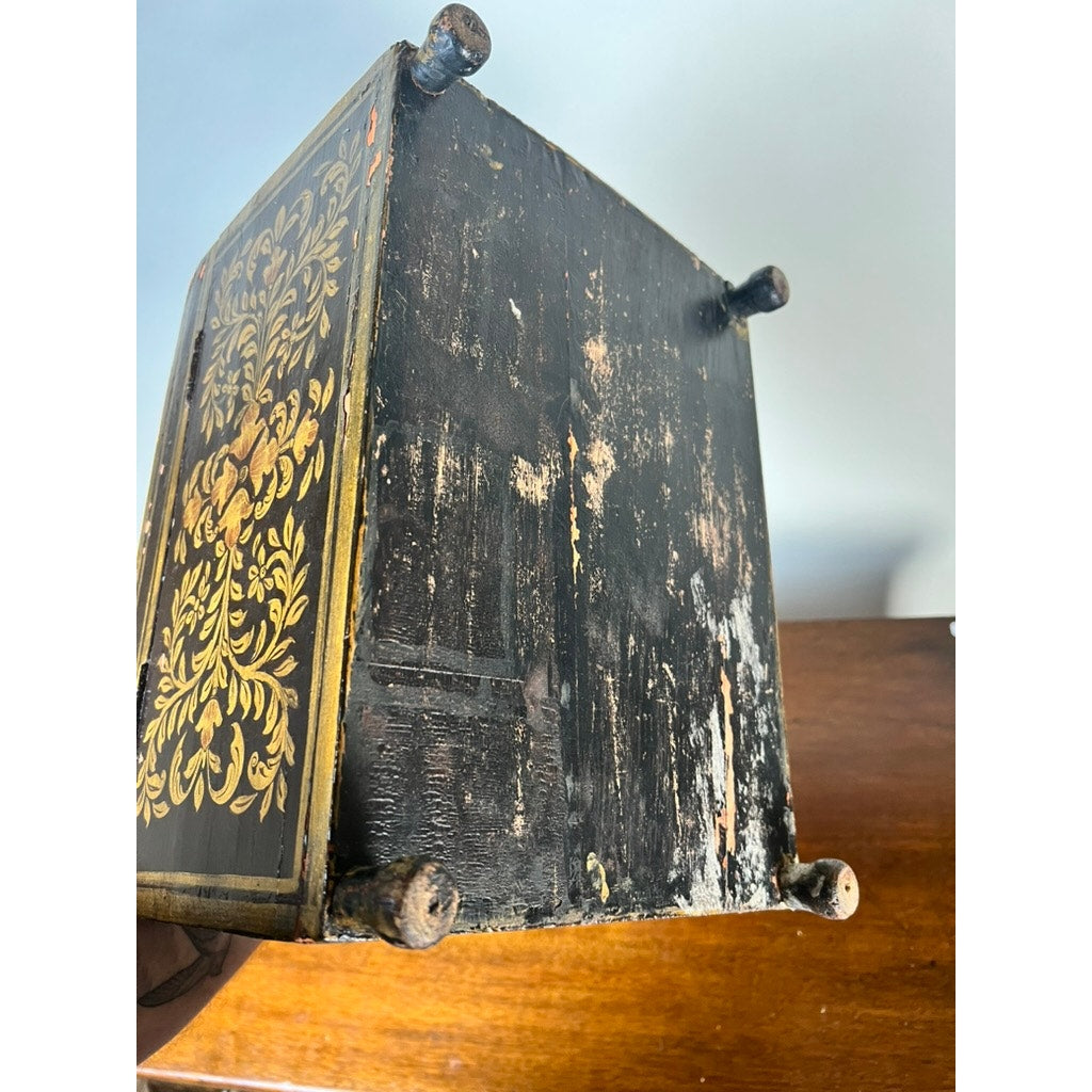 The photo shows the worn underside of an Antique New England Folk Art Painted Portrait Document Box (Mother & Child, c. 1840–1860), with chipped black paint, gold floral detail, four round feet, and exposed wood. Brand is unknown.