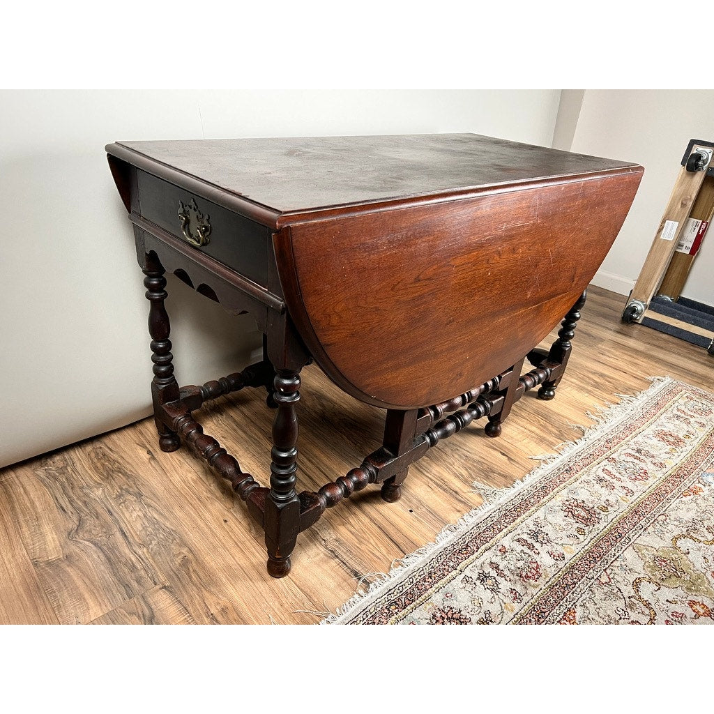 An Antique English William & Mary Walnut One Drawer Gateleg Dining Table (early 18th century) with dark turned legs and two drop leaves sits on a light wood floor next to a patterned rug, featuring a drawer with a brass handle.