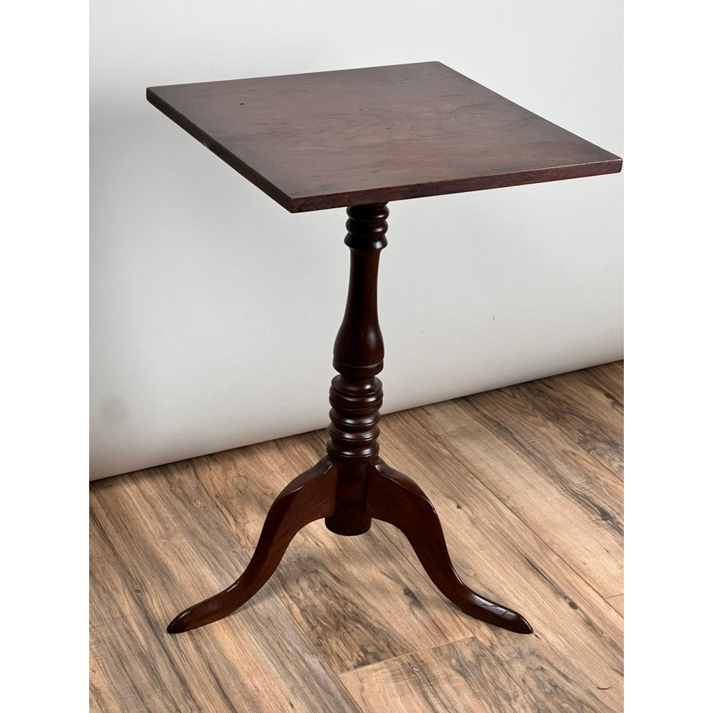 An Antique Low Cherry Candle Stand from the mid 19th century stands on a wooden floor against a white wall, featuring dark wood, a single turned pedestal leg, and three curved feet. Brand: None/Unknown.