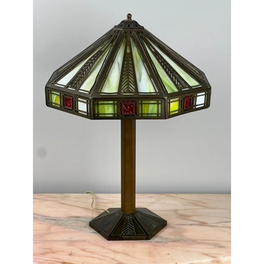 An Antique Bradley & Hubbard Arts & Crafts Prairie School Slag Glass Lamp with a geometric green, white, and red stained glass shade sits on a bronze-colored base atop a marble surface.
