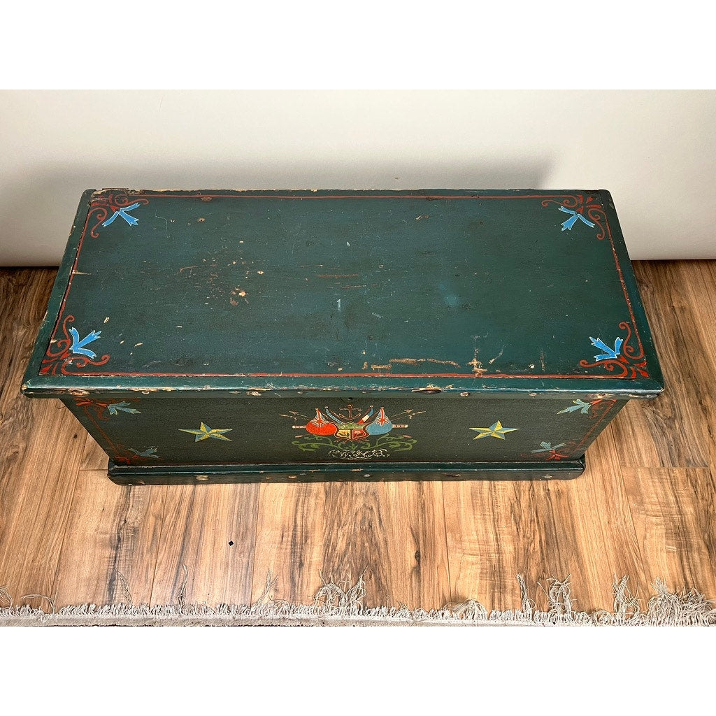 An Antique Paint Decorated English Sea Chest from the late 19th century, featuring red and blue birds, yellow stars, and a central motif, sits on a wooden floor beside a fringe-edged rug.