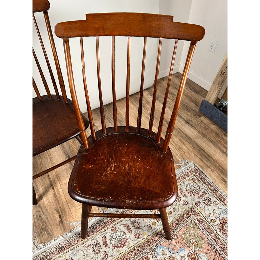 An Antique JR Hunt New Hampshire Step Down Windsor Chair from the early 19th century features a curved backrest and vertical slats, showing seat wear. It stands on a rug and wood floor next to another matching chair, part of a signed set of four.