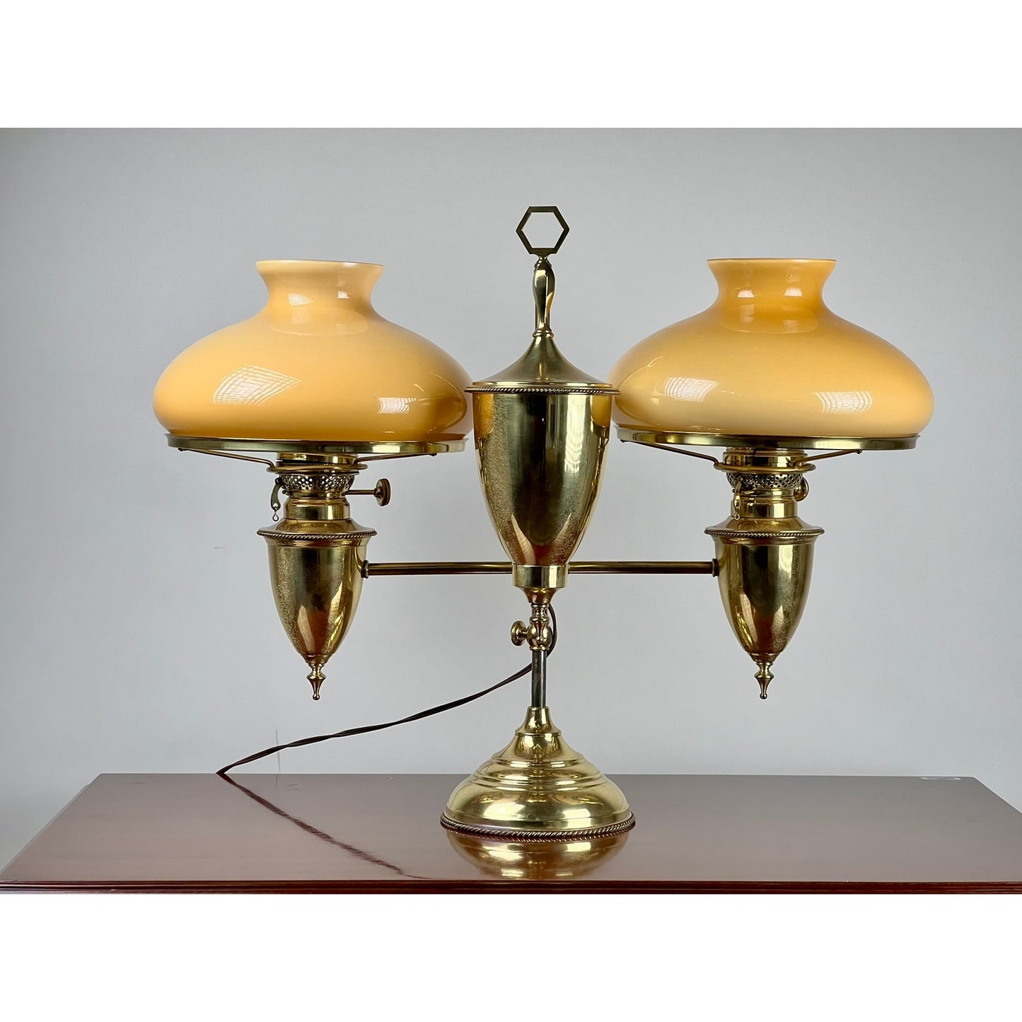 An Antique Bradley and Hubbard Brass Double-arm Student Lamp with caramel shades by Bradley and Hubbard is shown on a wooden surface against a plain gray background.