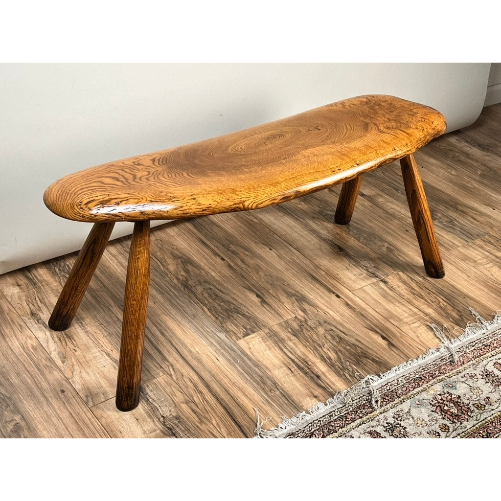 A Vintage Hand Made Oak Wabi Sabi Bench signed Neil Panyik sits on a wood-patterned floor beside a patterned rug.
