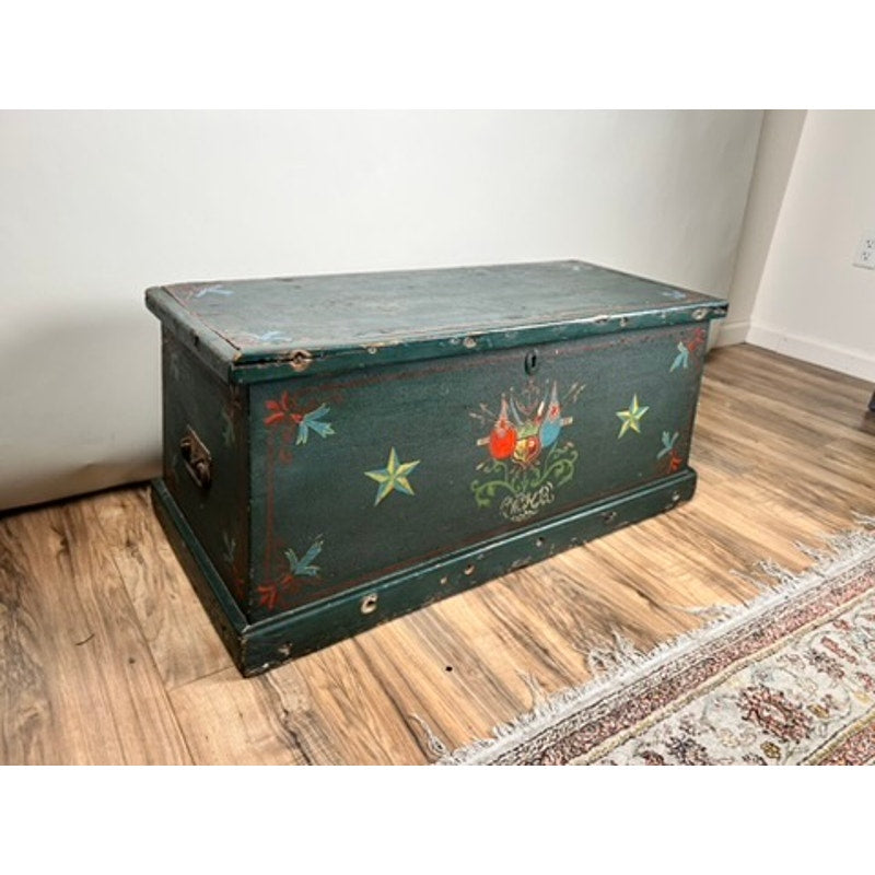 An Antique Paint Decorated English Sea Chest (late 19th century) with a dark green finish and colorful stars, flowers, and patterns sits on a wooden floor by a patterned rug against a plain white wall. Brand: None/Unknown.